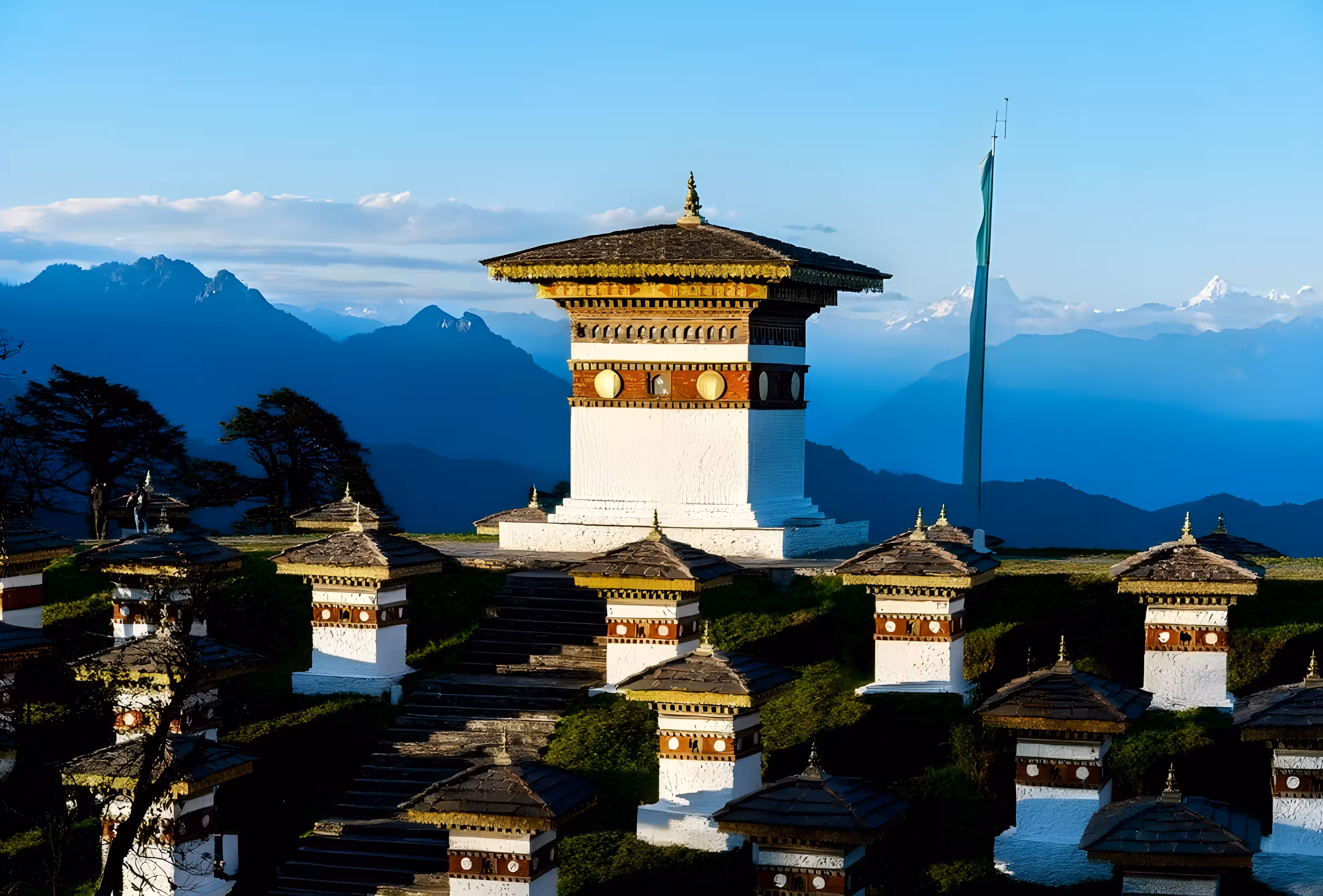 Panoramic view of Dochula Pass in Bhutan, showing snow-covered Himalayan mountains, 108 memorial chortens, and colorful prayer flags fluttering along the mountain pass road.